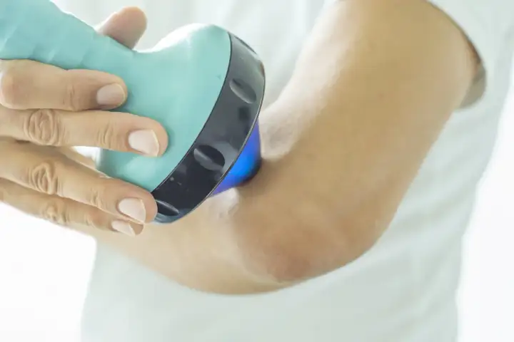 Une main utilise un appareil bleu clair pour soigner une tendinite au coude. La lumière bleue émise par l'appareil cible la zone douloureuse.